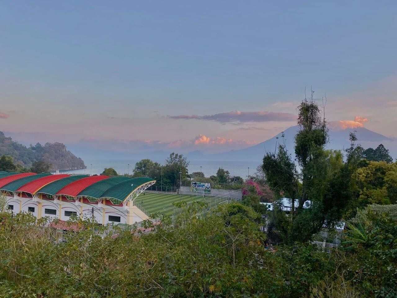 A stadium with a green roof, surrounded by trees and a mountain in the background.