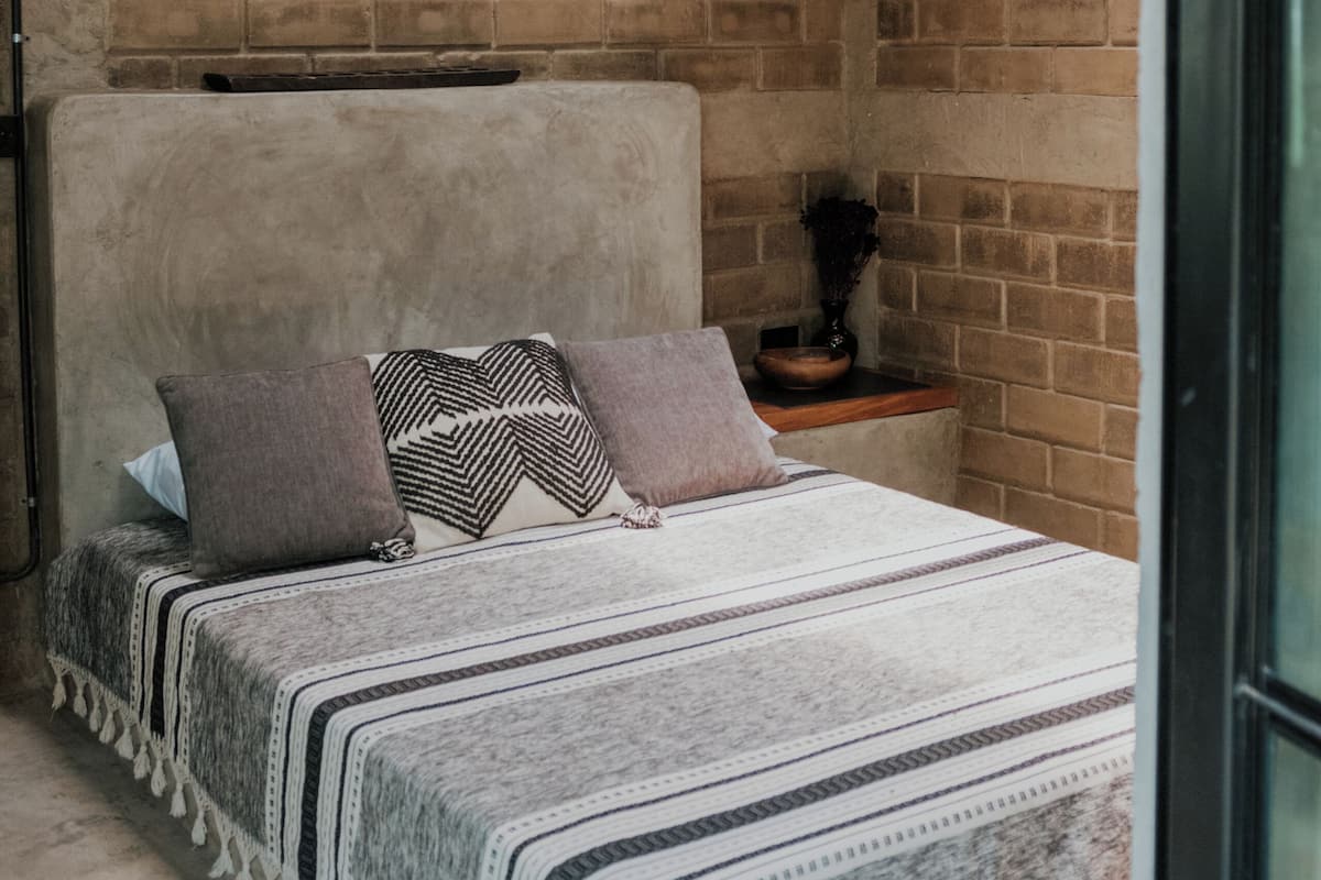 Ein Schlafzimmer mit Backsteinwand, einer gewobenen Leuchte, einem Bett mit gemusterten Bettwaren und einem Nachttisch mit einer Vase.