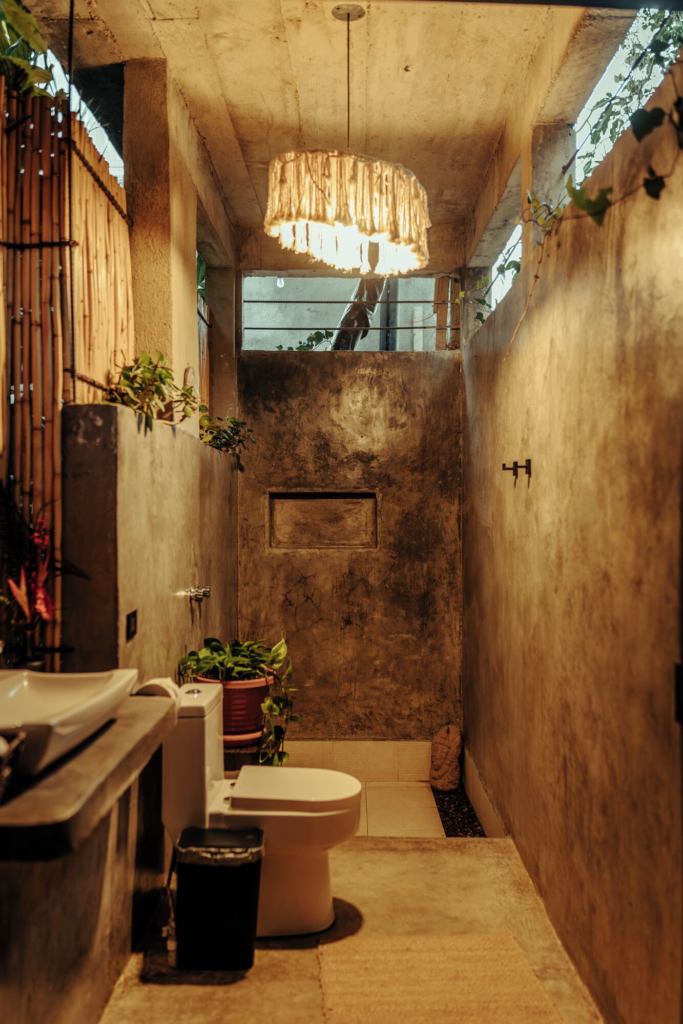 A narrow, concrete-walled bathroom with a toilet, sink, and plants.