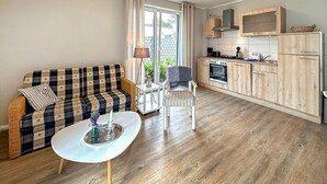 Ein kompakter Wohnbereich mit einer Couch, einem kleinen Tisch, einem Stuhl, einer Küchenzeile mit Holzschränken und einem Fenster mit Vorhängen.
