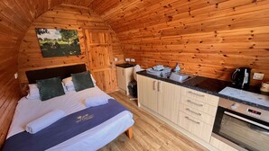 A cozy cabin interior with a wooden interior, a bed with pillows, a small kitchenette, and a painting on the wall. - Somerset glamping pod with access to adventure activities. (East Huntspill)