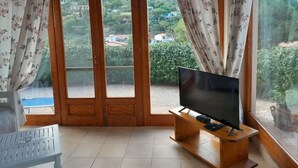 Une pièce avec un meuble TV en bois, une table blanche et une vue sur un paysage verdoyant, visible à travers une porte vitrée ornée de rideaux à motifs floraux.