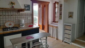 Une cuisine équipée d’une cuisinière à gaz, d’une machine à café et d’une petite table avec une chaise. Il y a une fenêtre avec des rideaux et une porte donnant sur l’extérieur.
