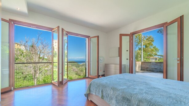 Une chambre dotée d’un grand lit, d’une porte-fenêtre coulissante donnant sur un balcon, et offrant une vue sur la verdure et les bâtiments.