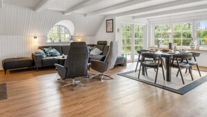 En moderne loftlejlighed med højt til loftet, et hyggeligt opholdsområde med sofa og stole, et spisebord med stole og store vinduer med udsigt til grønne omgivelser.