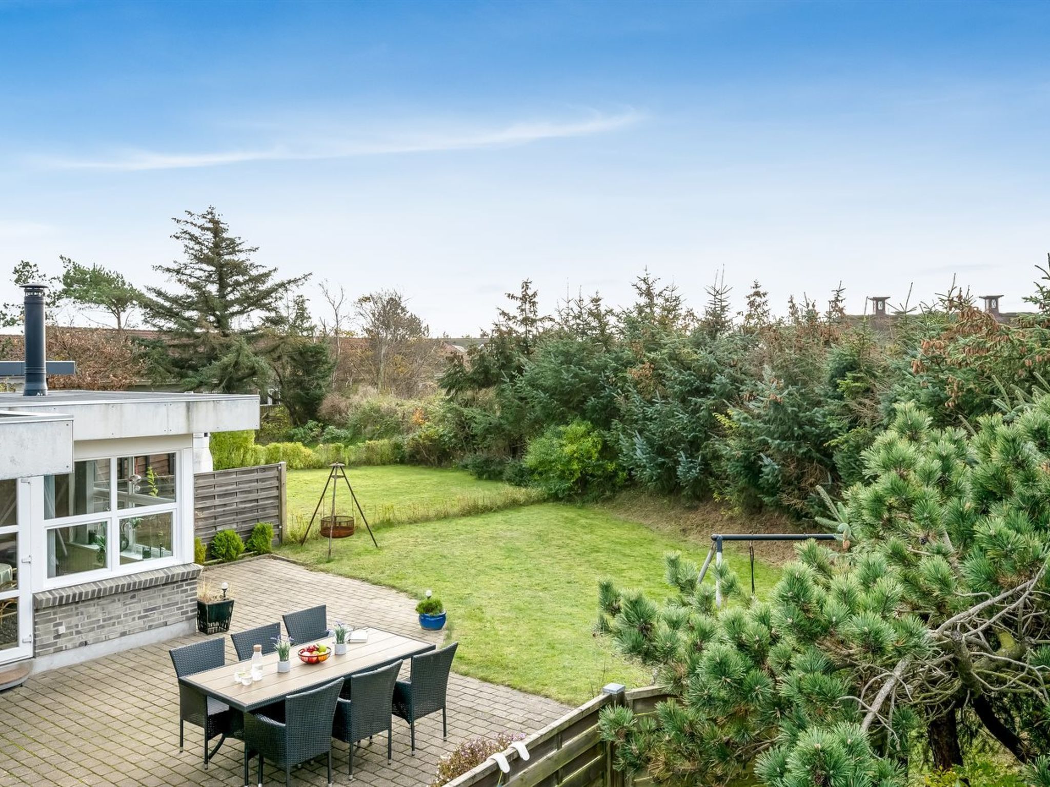 Une maison moderne dotée d’un patio comprenant une table et des chaises, le tout entouré d’une pelouse bien entretenue et d’une végétation dense.
