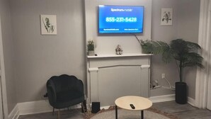 A modern room with a mounted television displaying a Spectrum mobile advertisement, a fireplace, a black chair, a small round table, and a potted plant. - 2 bed room Apartment (Albany)