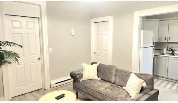 A living room with a grey sofa, a small round table, and a remote control. There are two white doors and a kitchen area with cabinets and appliances. - 2 bed room Apartment (Albany)