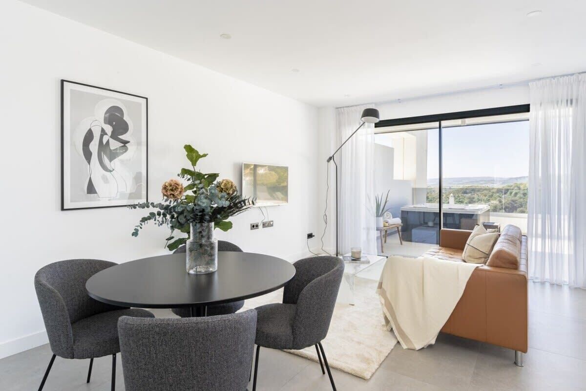 Un salon moderne avec une table à manger ronde, des chaises recouvertes d’un tissu gris, un canapé agrémenté d’un plaid blanc et une grande fenêtre donnant sur la ville.