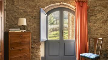 Una stanza con parete in pietra, un comò in legno con una lampada, una sedia e una finestra con tende.