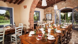 Un'area pranzo con un lungo tavolo in legno apparecchiato per un pasto, circondata da archi in mattoni e travi in legno.