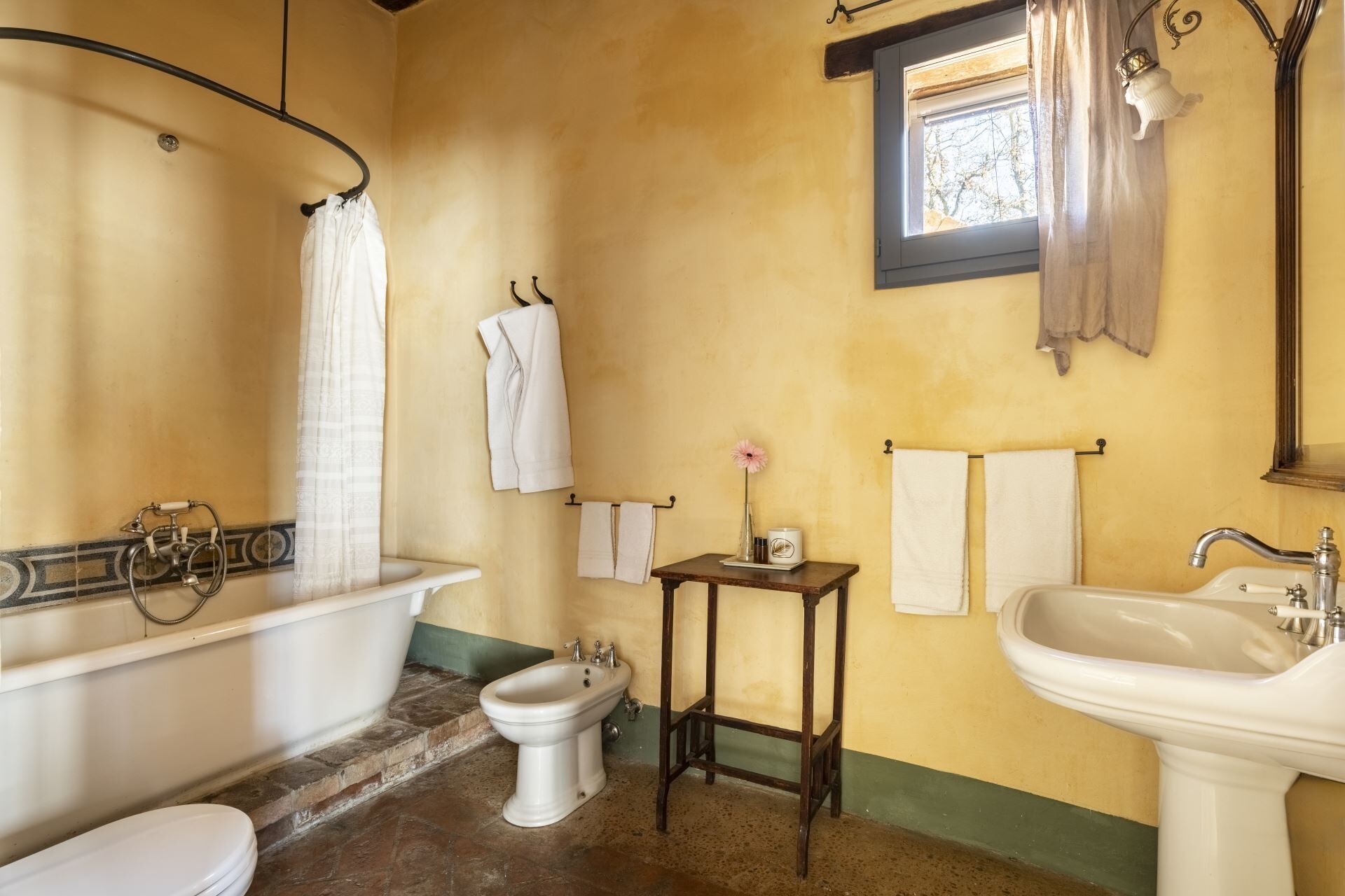 A bathroom with a bathtub, toilet, bidet, and sink. There is a small table with a vase and a mirror above the sink.