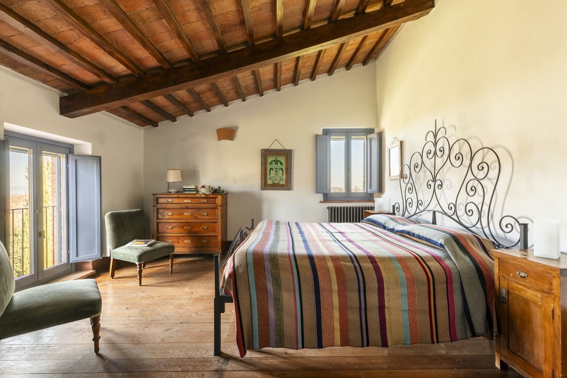 A bedroom with a wooden ceiling, a bed with a striped blanket, a dresser, a chair, and a window with shutters.