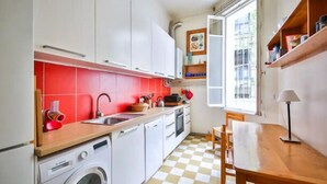 Een kleine keuken met witte kastjes, een rode spatwand, een geruit vloertje en een raam voor natuurlijk licht.