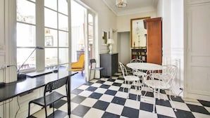 Ein Zimmer mit Schachbrettmuster-Boden, weißen Wänden, einem runden Tisch mit weißen Stühlen, einem schwarzen Schreibtisch mit einem Stuhl und einer Holztür.