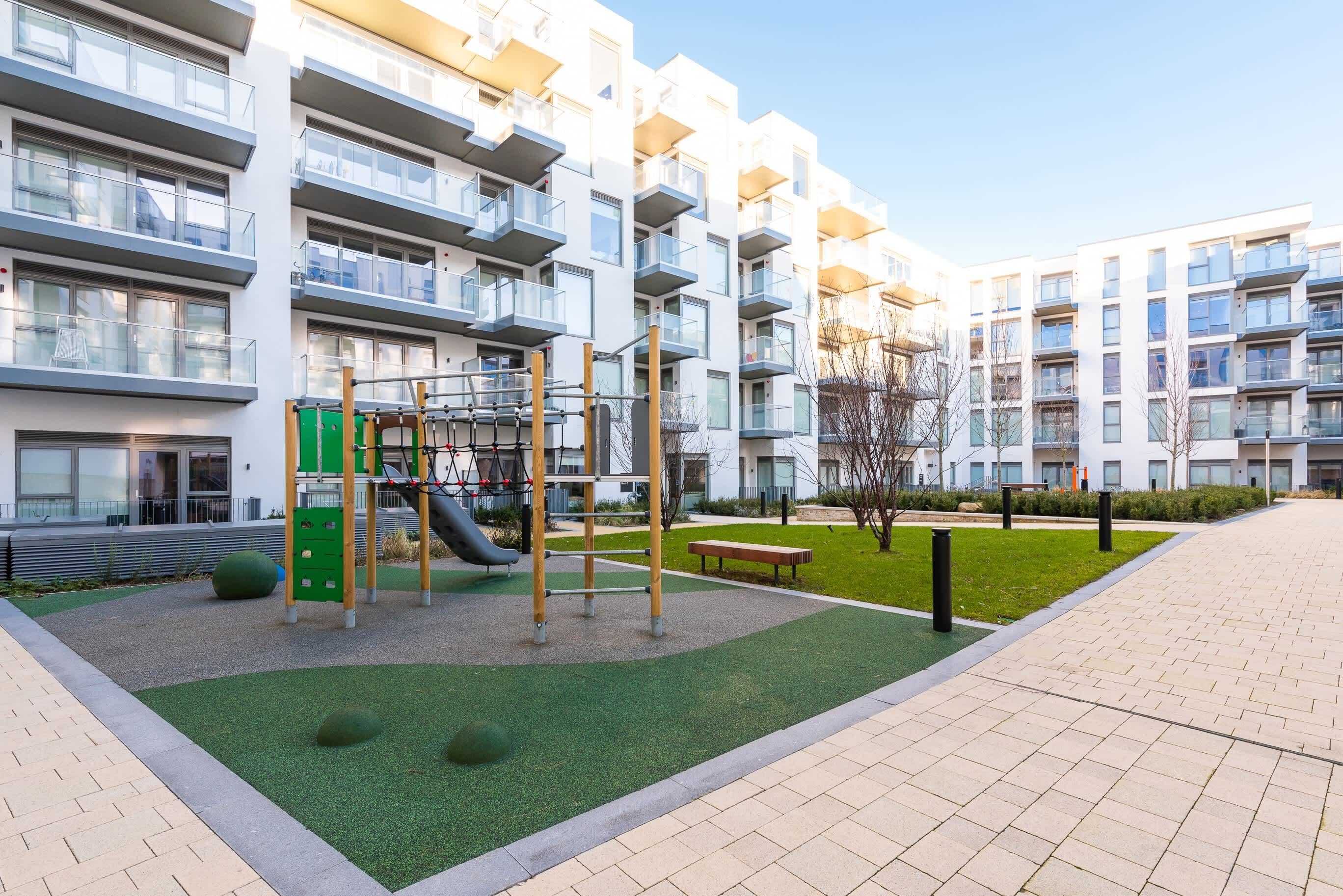 A playground with a slide and climbing structures, surrounded by a landscaped area with grass and trees, in front of a modern apartment building.