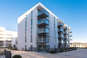Un immeuble moderne à plusieurs étages, doté de balcons, d’une cour dallée et surplombant un ciel dégagé.
