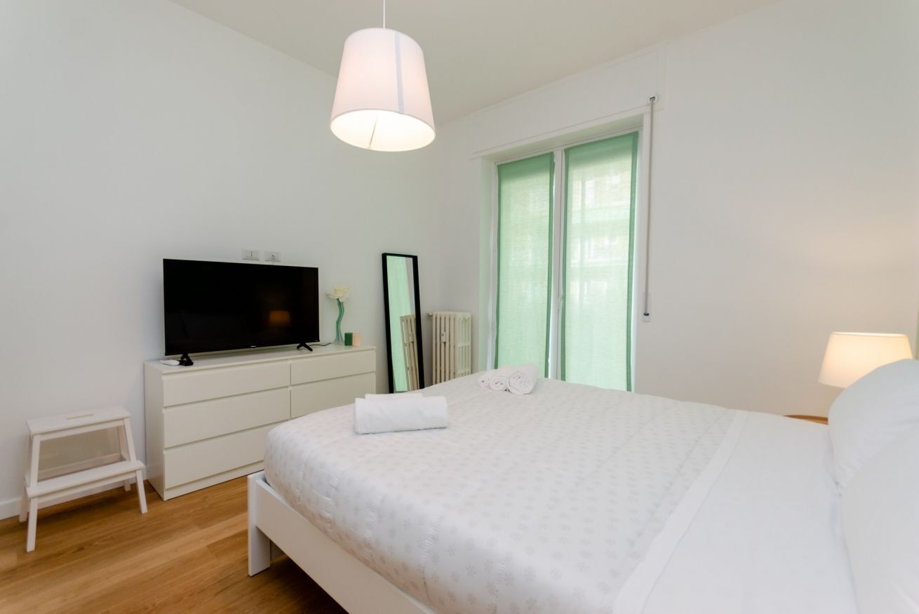Une chambre à coucher avec un lit, un téléviseur posé sur une commode, une petite table et une fenêtre avec des rideaux.