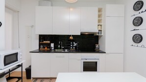 Una cocina moderna con armarios blancos, encimera negra, microondas, fregadero y horno pequeño. Hay relojes en la pared y una lámpara en el techo.
