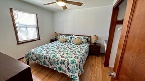 A bedroom with a bed, two nightstands, a ceiling fan, a window with blinds, and a wooden door. - One Particular Cottage - Next to Margaritaville (Fort Myers Beach)