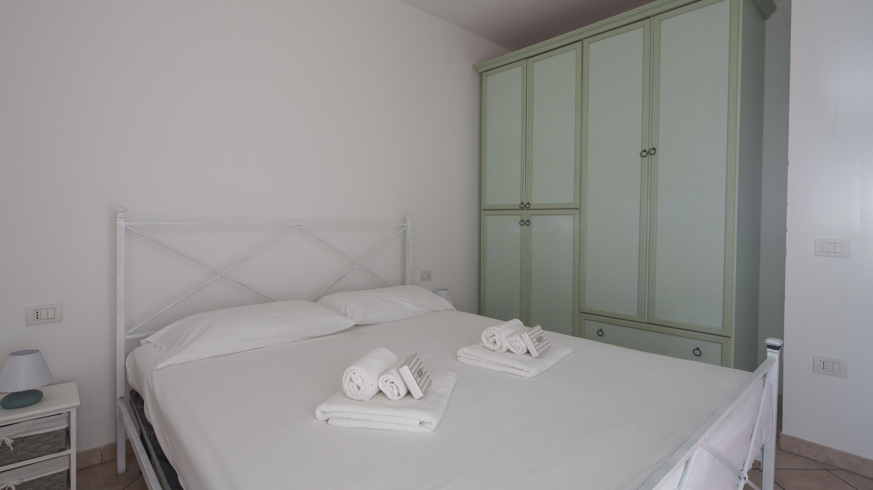 A neatly made bed with white linens and towels, a white bedside table with a lamp, and a green wardrobe with two doors.