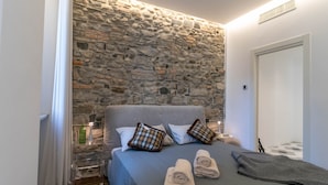 Une chambre avec un mur en pierre, un lit avec deux oreillers, une table de chevet avec une lampe et une porte.