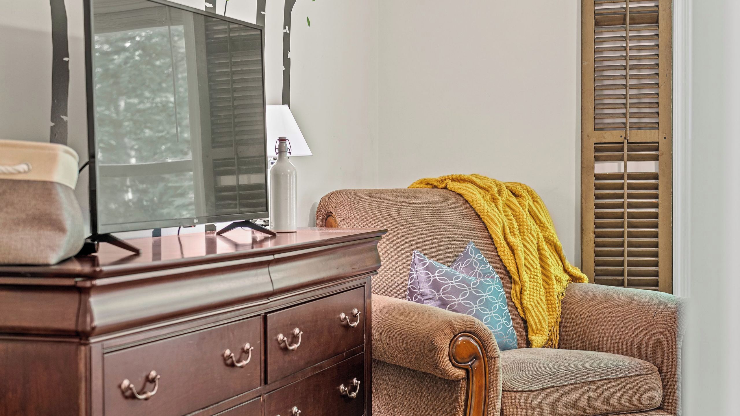 A living room with a wooden dresser, a television, a sofa with a yellow throw blanket, and a window with blinds.