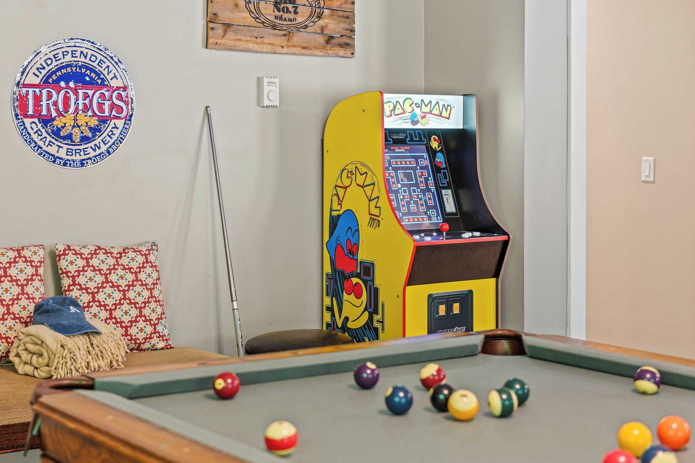 A pool table with various colored balls, a pool cue, and a Pac-Man arcade machine.