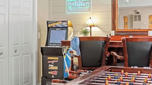 A room with a foosball table, a retro arcade machine, a neon sign for a tavern, and a mirror reflecting a cabinet and speakers.