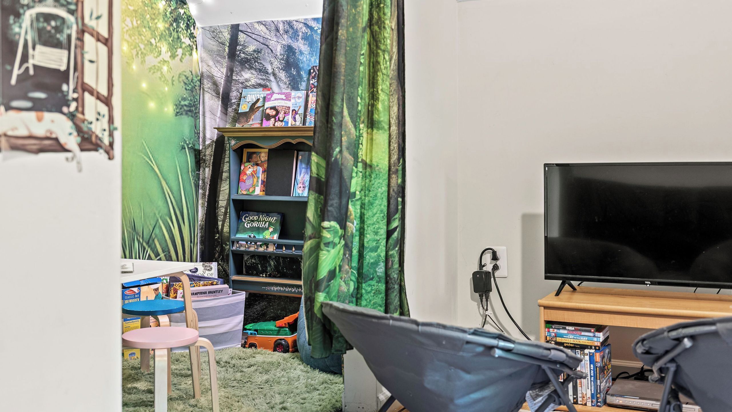 A room with a TV, a bookshelf with books, a small table, and a chair.