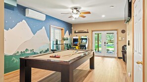 A pool table with pool cues and balls, a ceiling fan, air conditioning unit, and arcade games in a room with wooden flooring and a mountain mural.