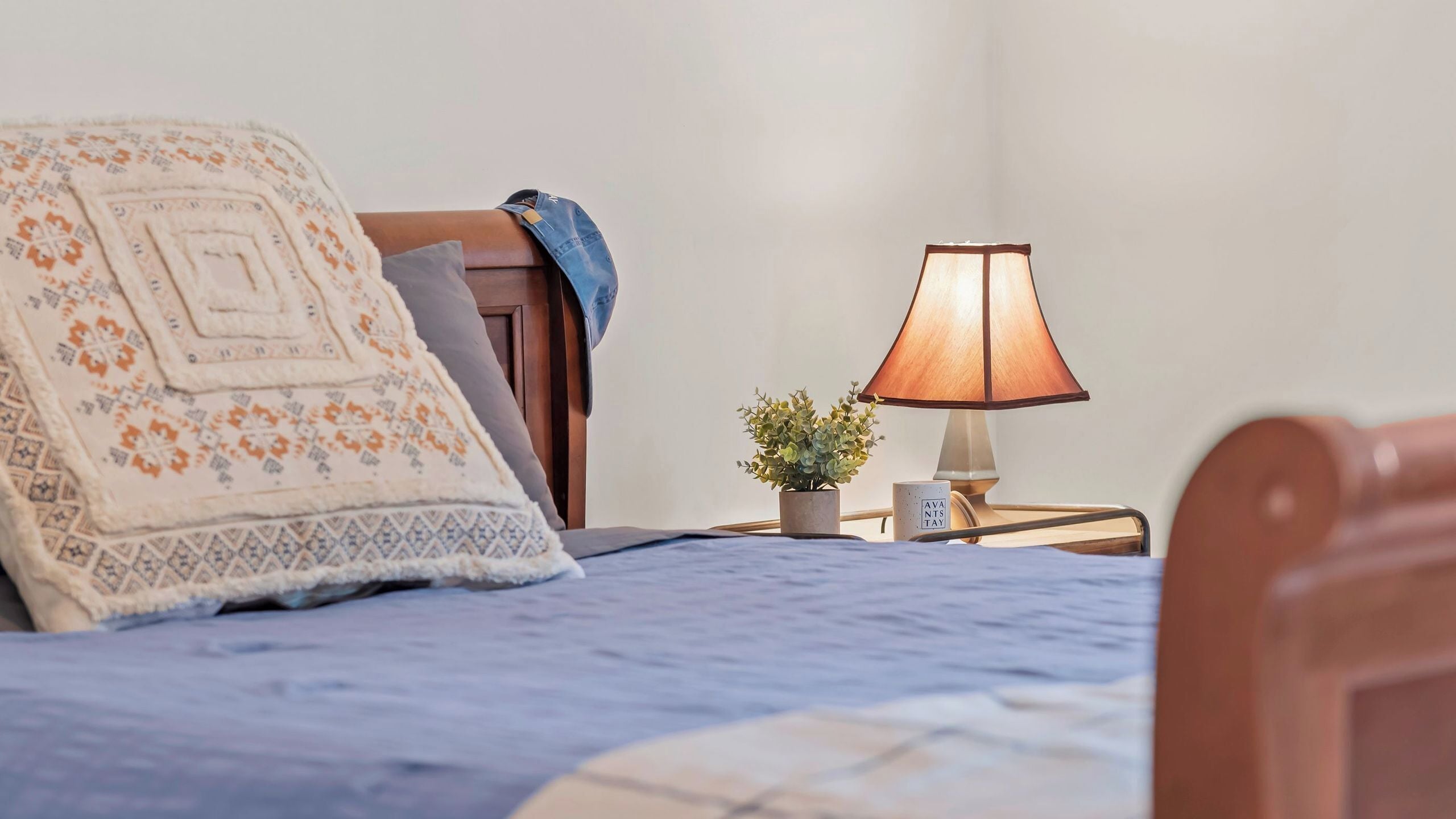 A neatly made bed with a patterned blanket, a bedside lamp, a small potted plant, and a wooden headboard.