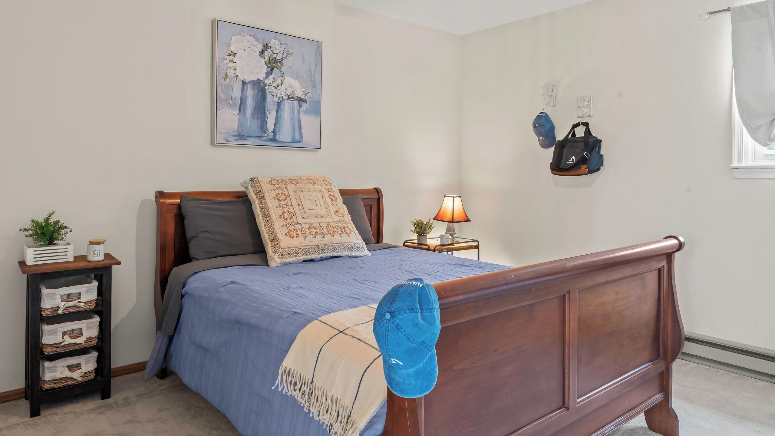 A bedroom with a wooden bed, a nightstand, a lamp, a wall-mounted bag rack, and a painting.