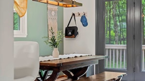 A dining area with a wooden table, a white chair, a vase with a plant, a hanging bag, and a view of a deck through glass doors.