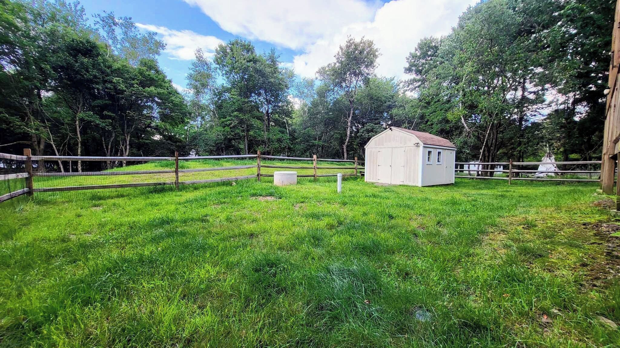 Un espace clos délimité par une clôture, avec un petit bâtiment blanc, une palissade en bois et des arbres en arrière-plan.