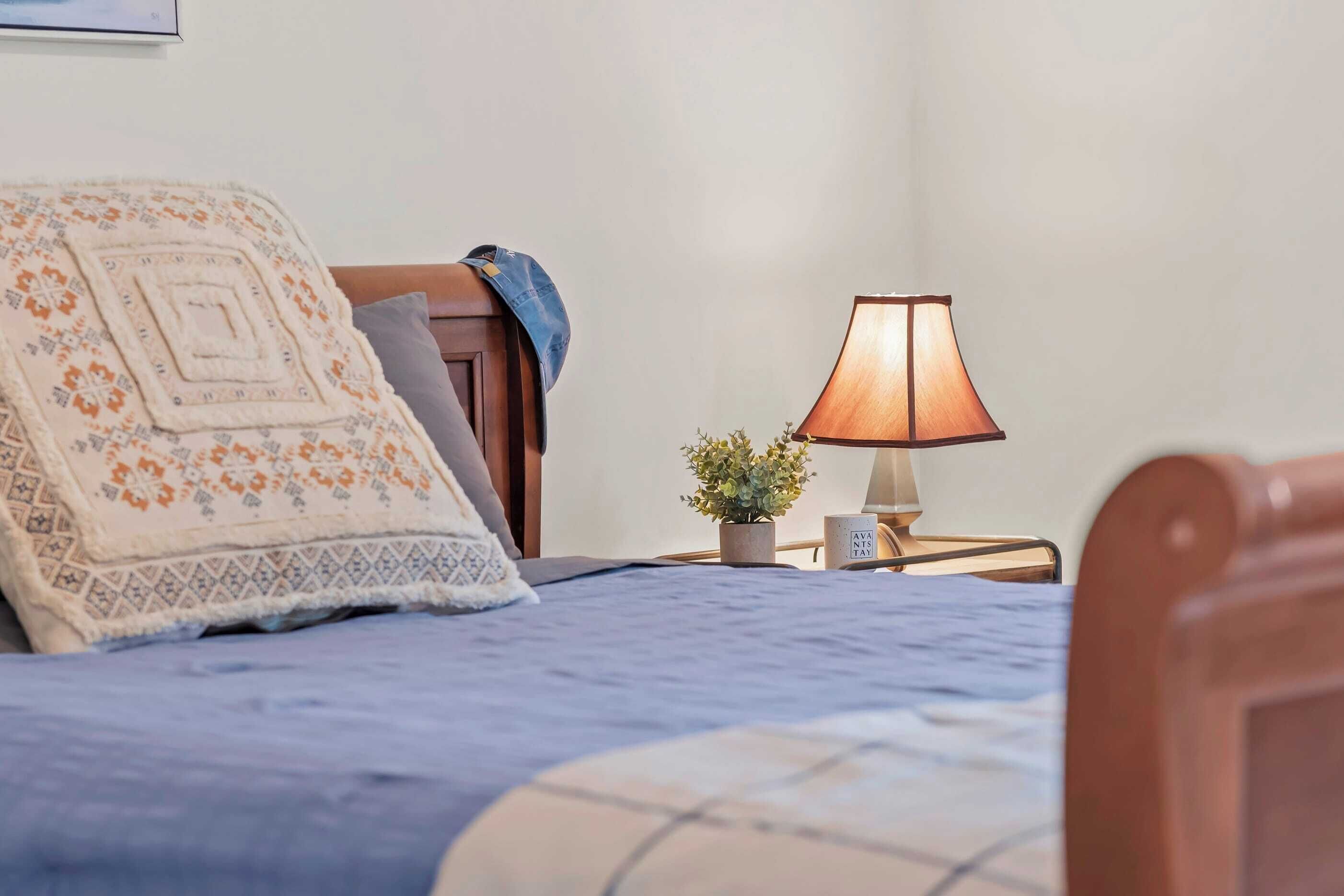 Ein ordentlich gemachtes Bett mit blauer Decke und einem dekorativen Kissen, ein Nachttisch mit Lampe und einem kleinen Topfpflanzen sowie ein Holzkopfstück.