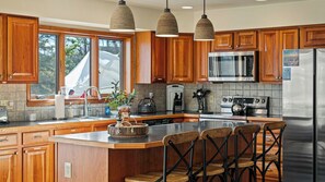 A modern kitchen with wooden cabinets, a large island with bar stools, a stainless steel refrigerator, and a microwave. - Entertainer's Home w/ Deck, Game Room & Hot Tub Lake View Retreat by Avantstay (Albrightsville)