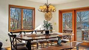 A dining area with a large wooden table, multiple chairs, a chandelier, and large windows with a view of trees and a grill outside. - Entertainer's Home w/ Deck, Game Room & Hot Tub Lake View Retreat by Avantstay (Albrightsville)