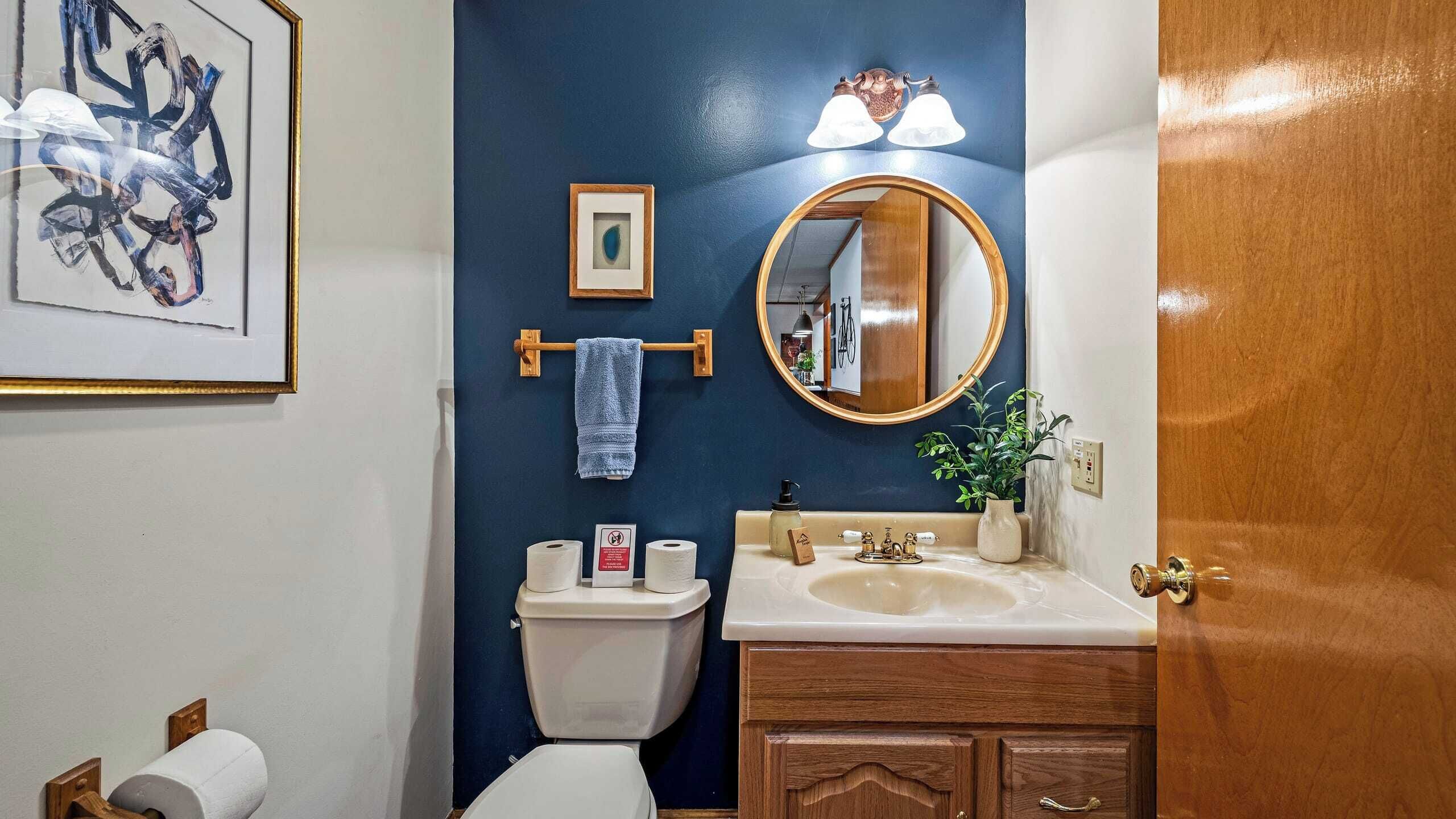 A bathroom with a blue accent wall, a toilet, a sink with a mirror, and a framed picture above the toilet.