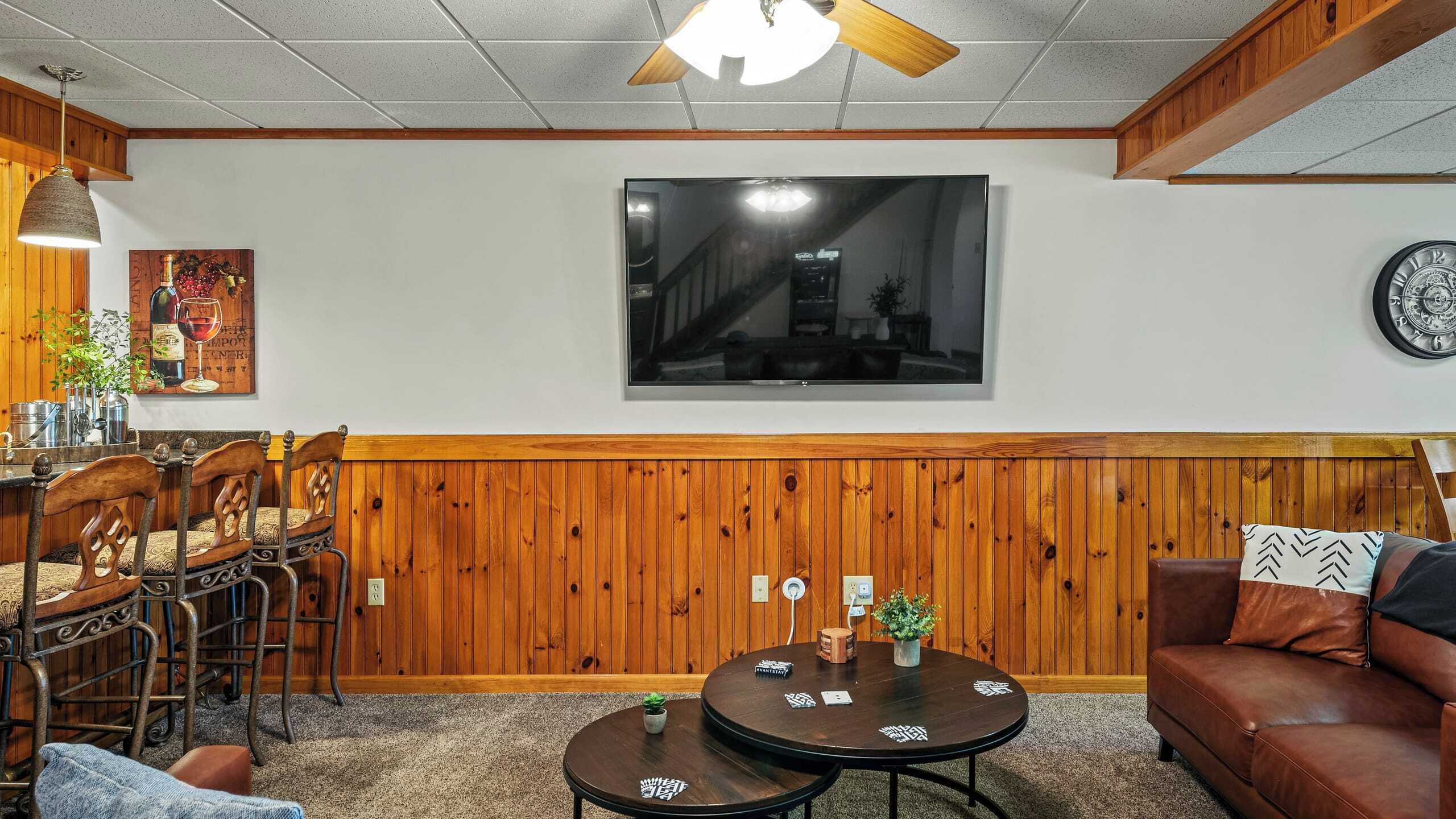 Een gezellige kamer met houten lambrisering, een grote flatscreen televisie aan de muur, een zithoek met stoelen en een tafel, en een leren bank.