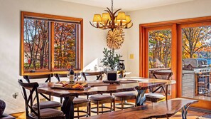 A dining area with a large wooden table, chairs, a chandelier, and a view of autumn trees through large windows. - Entertainer's Home w/ Deck, Game Room & Hot Tub Lake View Retreat by Avantstay (Albrightsville)