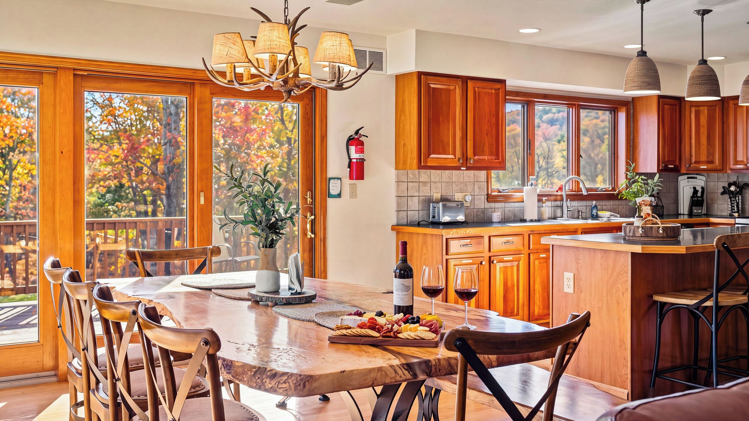 A kitchen with wooden cabinets, a central island with a wine bottle and glasses, and a view of autumn trees through large windows.