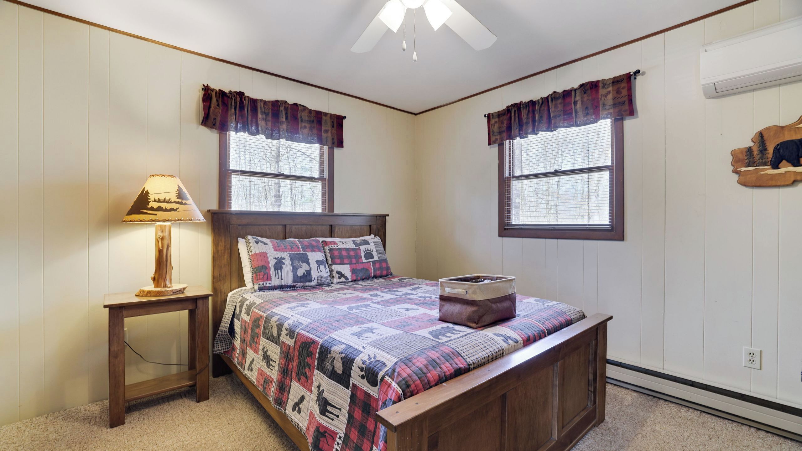 A bedroom with a wooden bed, two windows with curtains, a nightstand with a lamp, and a wall-mounted air conditioner.