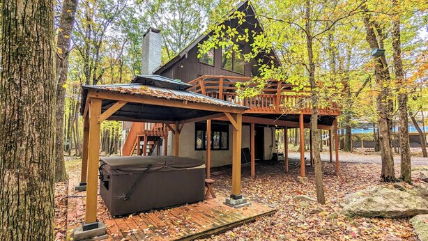 A wooden cabin with a hot tub and a deck, surrounded by trees and fallen leaves. - Hot Tub + Sauna + Deck + Firepit + Beach Access + Near Dining Grateful Escape by Avantstay (Lake Harmony)