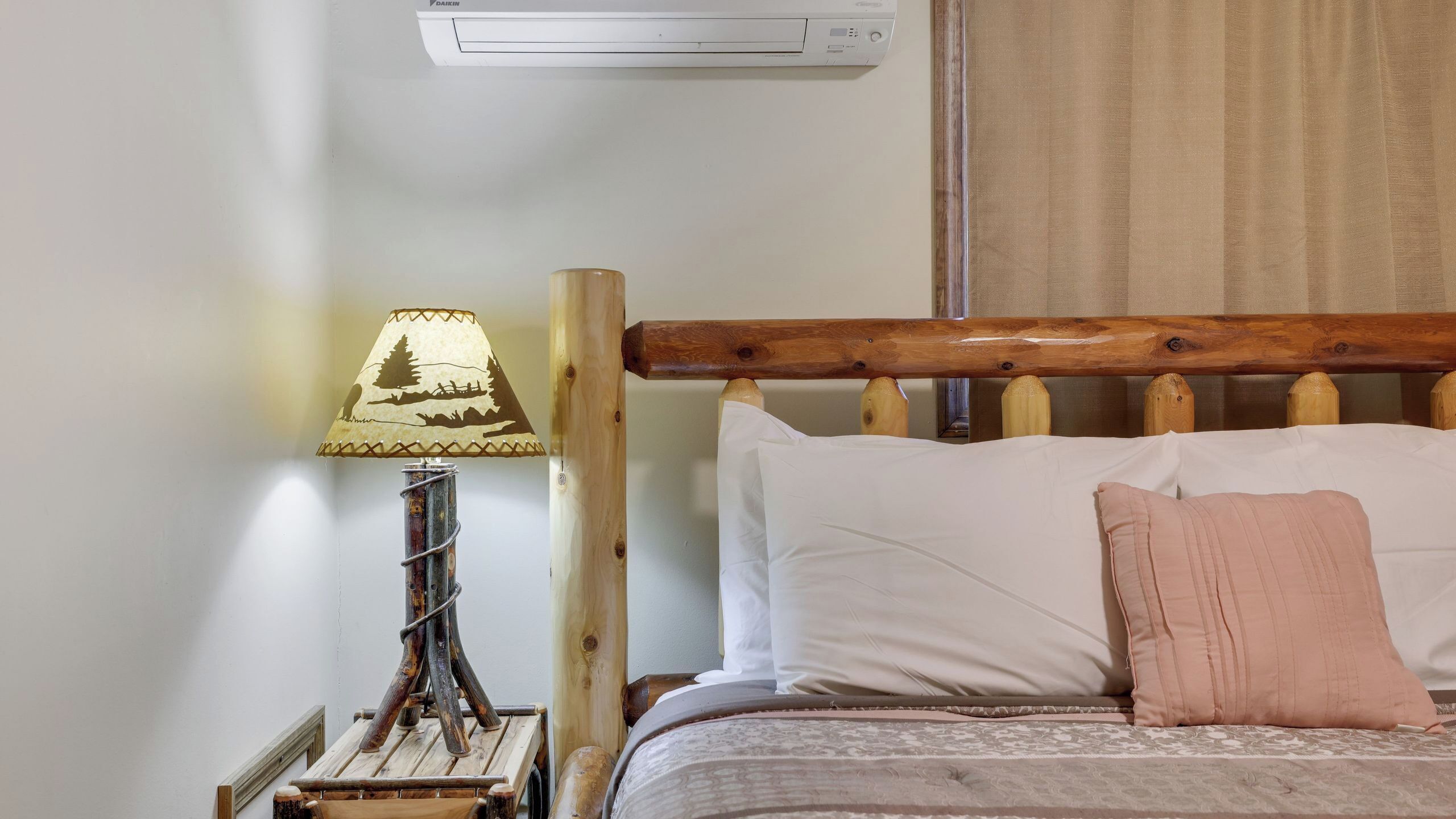 A bed with white and pink pillows, a wooden headboard, a lamp with a patterned shade, and a wall-mounted air conditioner.