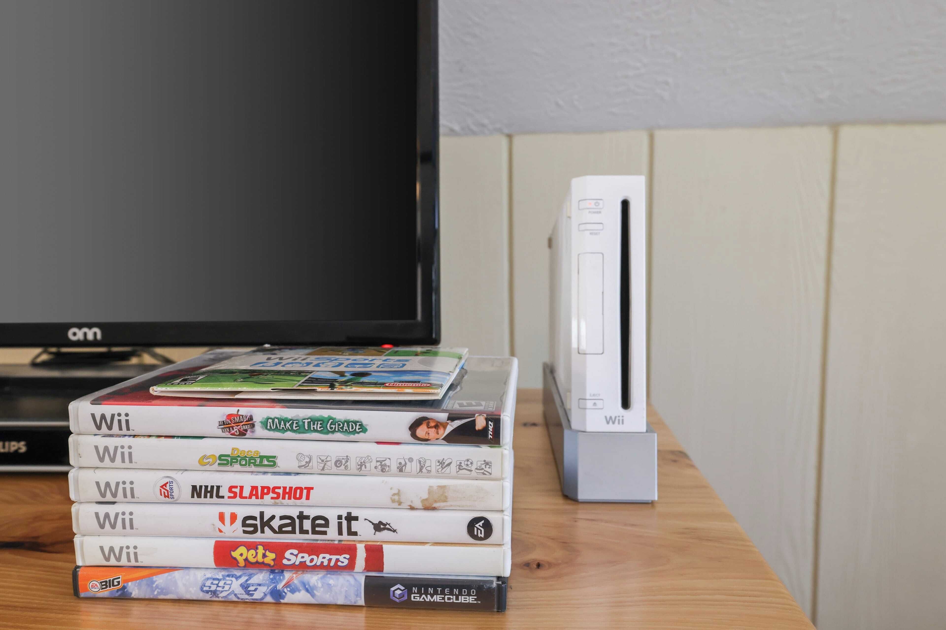 Un montón de cajas de juegos de Wii sobre una superficie de madera, con una consola Wii y un televisor al fondo.