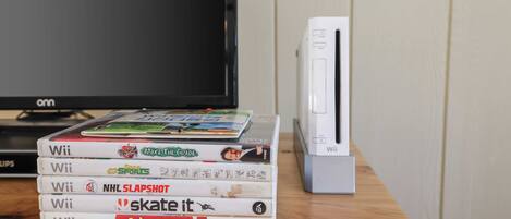 Un montón de cajas de juegos de Wii sobre una superficie de madera, con una consola Wii y un televisor al fondo.