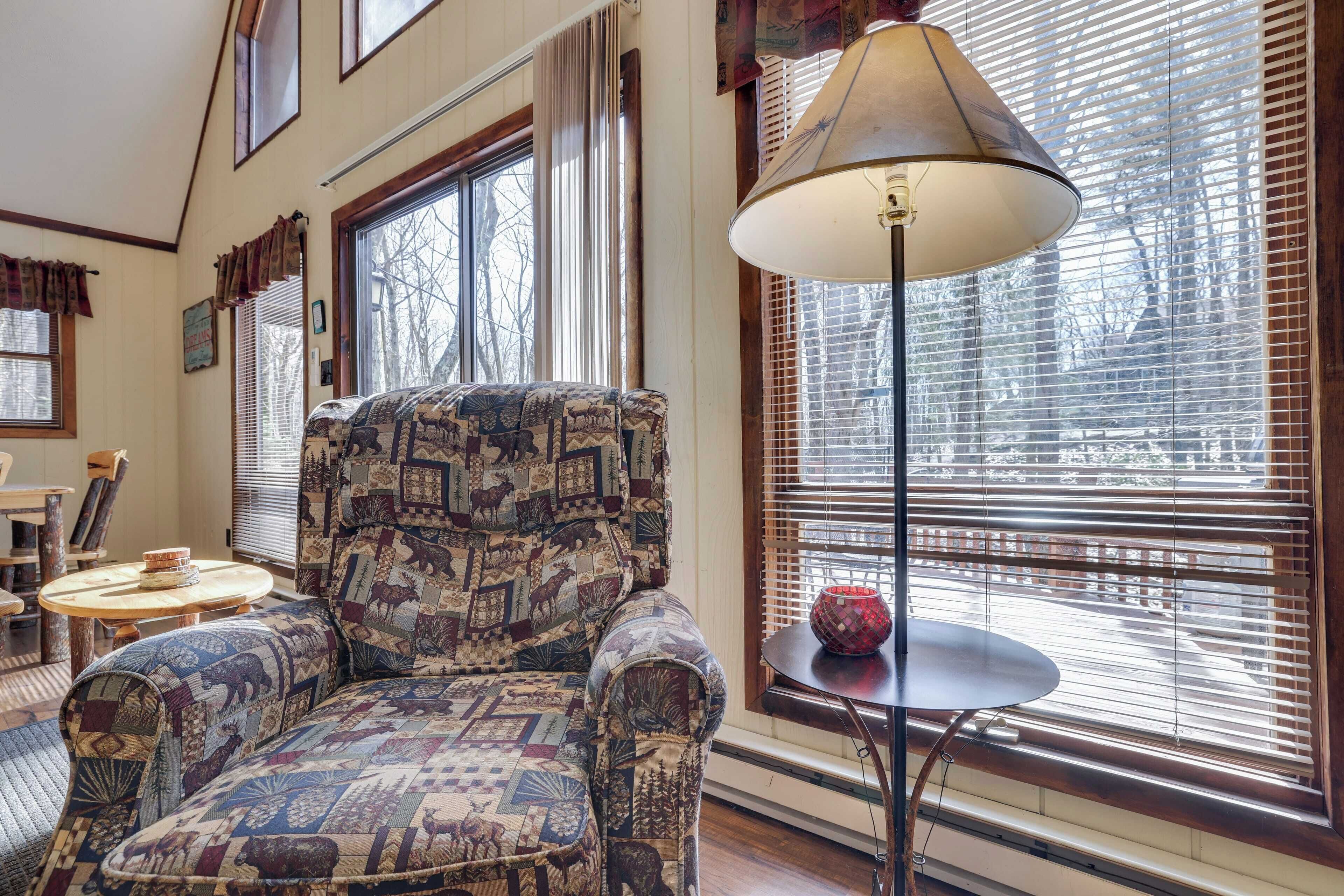 A cozy living room with a patterned armchair, a round table with a lamp, and large windows with blinds.
