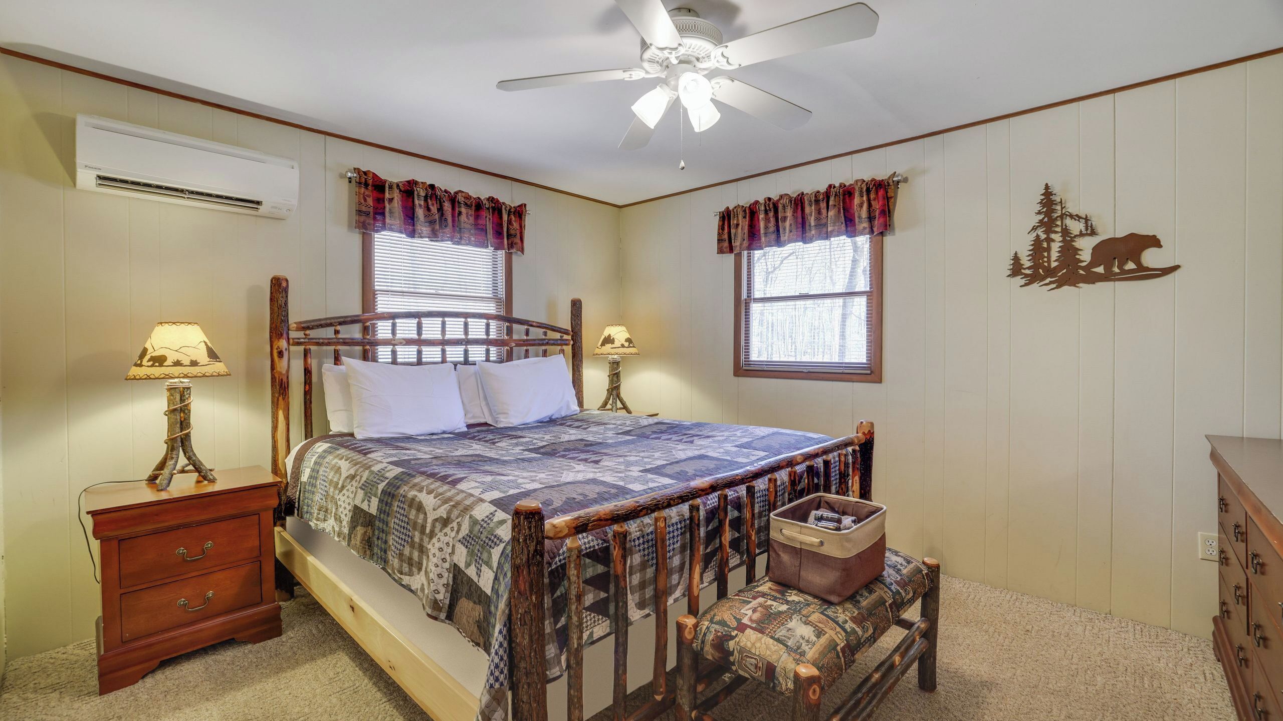 Une chambre avec un lit en bois, deux tables de chevet avec des lampes, un ventilateur de plafond, une fenêtre avec des rideaux et une décoration représentant un ours sur le mur.