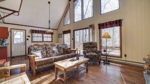 A spacious living room with a wooden floor, a sofa with patterned cushions, a coffee table, and a chair. There are large windows with curtains, a lamp, and a door leading outside. - Hot Tub + Sauna + Deck + Firepit + Beach Access + Near Dining Grateful Escape by Avantstay (Lake Harmony)